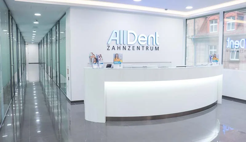 Empfangstresen in modernem Zahnarztzentrum mit beleuchtetem Schriftzug 'AllDent ZAHNZENTRUM' an weißer Wand und Glaswänden im Flur.