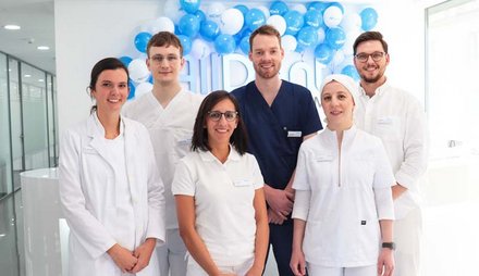 Sechs Personen in medizinischer Arbeitskleidung stehen vor einer Wand mit blauen und wei&szlig;en Luftballons.