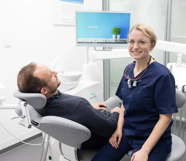 Eine Zahnärztin in blauer Arbeitskleidung sitzt neben einem Patienten der im Behandlungsstuhl aitzt und lächelt in die Kamera, im Hintergrund ist ein Monitor zu sehen